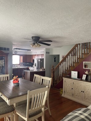 A dining area with a wooden table and chairs, a kitchen in the background, and a staircase leading to another level. - Waconda Roost (Glen Elder)