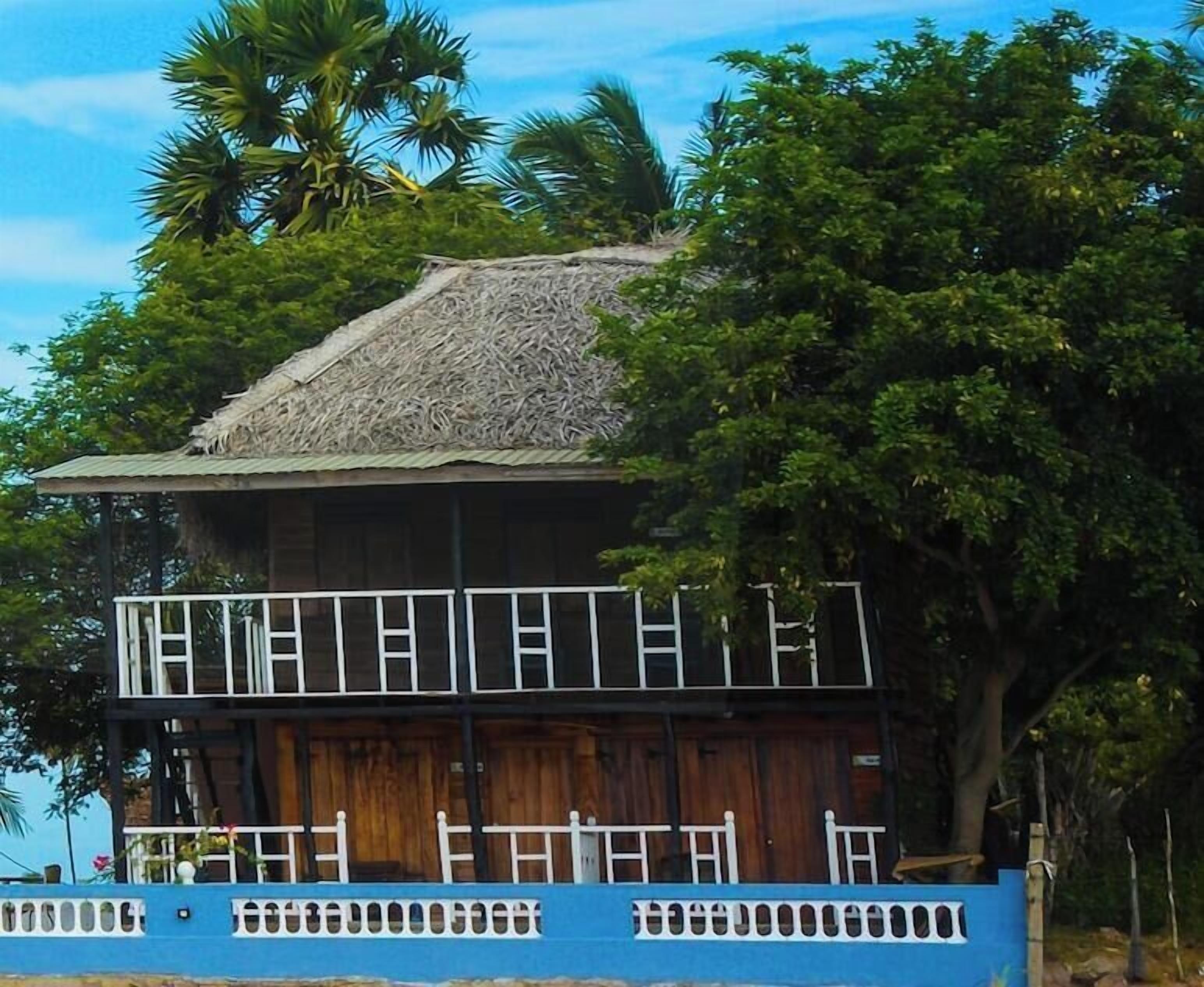 A two-story wooden building with a thatched roof, surrounded by lush greenery.