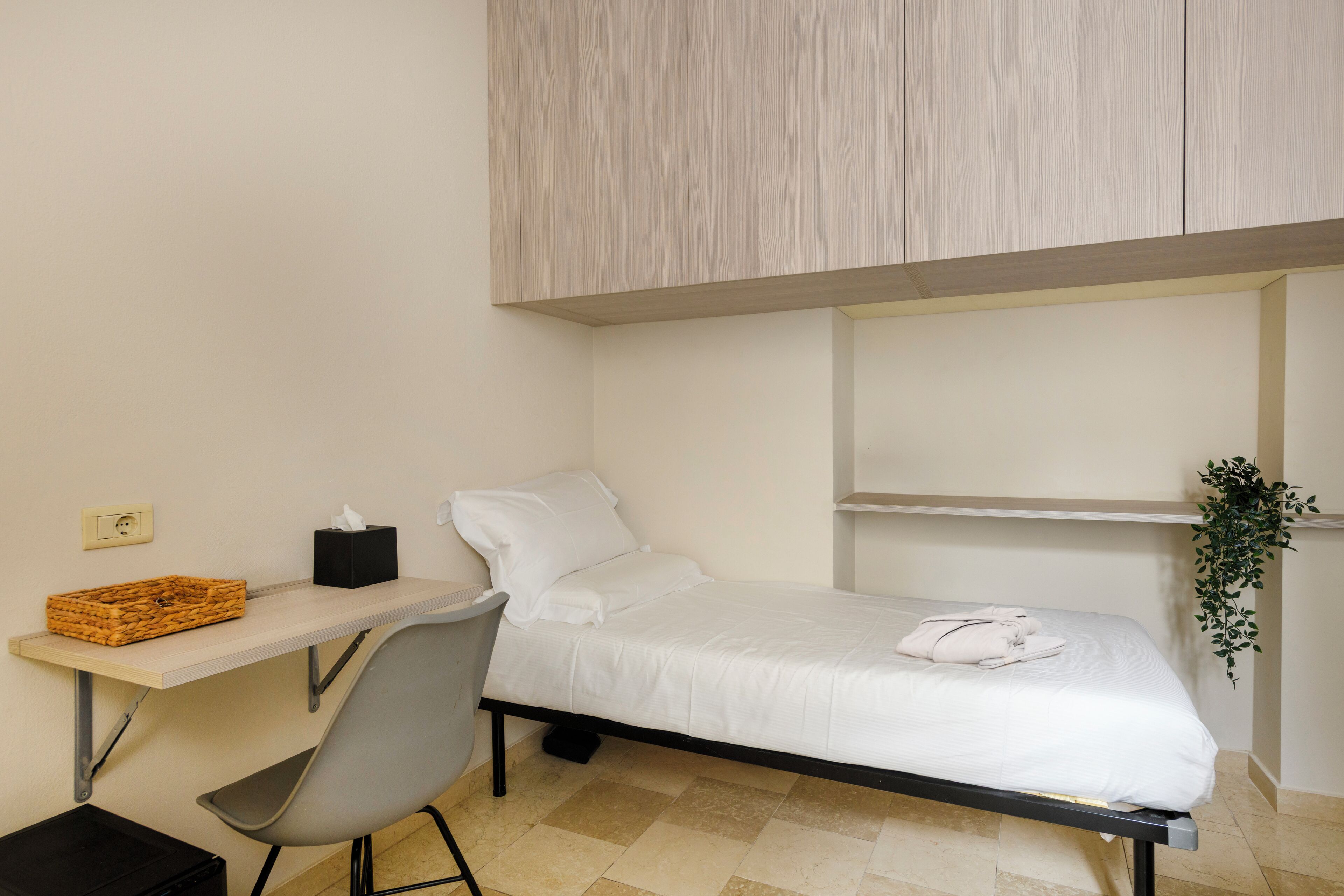 Ein kleiner, ordentlicher Raum mit einem Bett, einem Schreibtisch und einem Stuhl. An der Wand befindet sich eine Pflanze, auf dem Schreibtisch ein Korb und auf dem Boden eine schwarze Tasche.