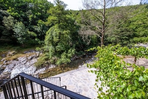 Ein Steinpfad führt zu einer Steinsbrücke über einen Fluss, umgeben von üppiger Vegetation und Bäumen.
