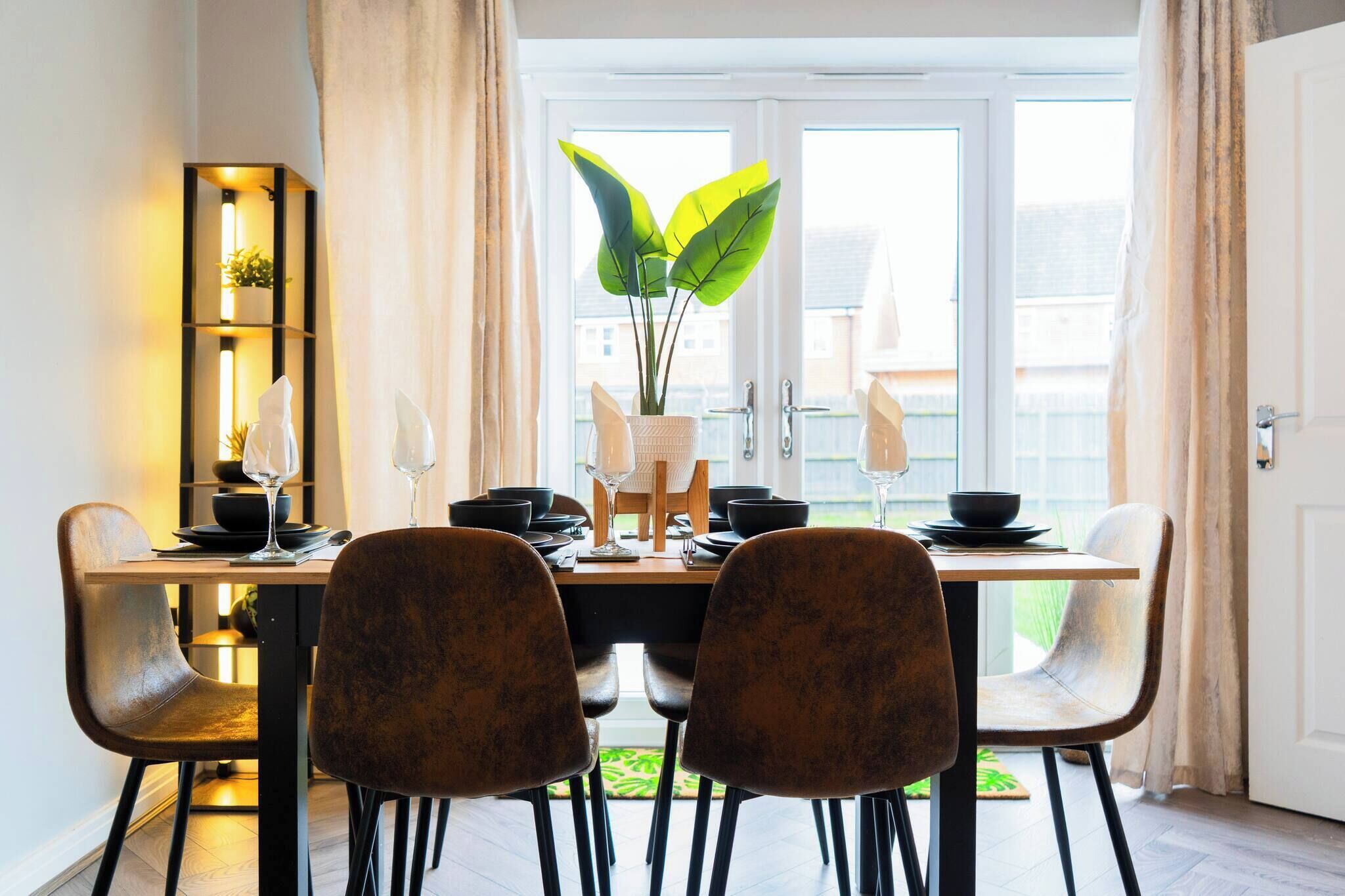 A dining area with a wooden table set for four, surrounded by upholstered chairs. A tall plant is centered on the table, and a modern, illuminated shelf unit is to the left.