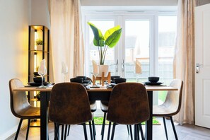 A dining area with a wooden table set for four, surrounded by upholstered chairs. A tall plant is centered on the table, and a modern, illuminated shelf unit is to the left. - Luxe 3BR - 8 Min to Town Centre - Bradgate Park & Leicester - Parking - Garden (Loughborough)