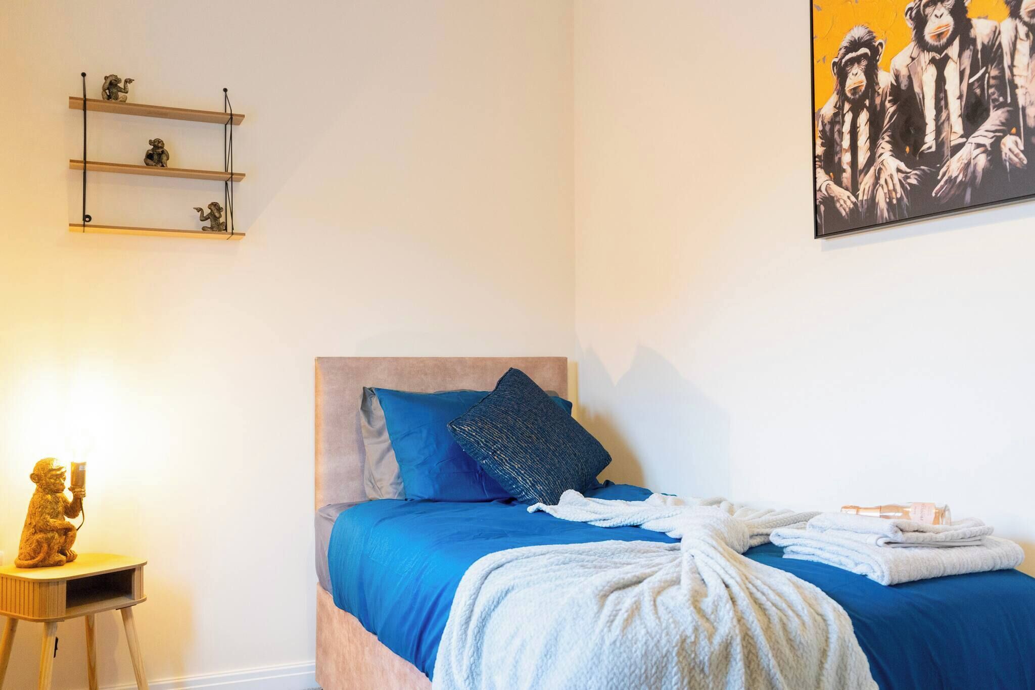 A bedroom with a bed, blue bedding, a white blanket, a small wooden nightstand with a statue, and a wall-mounted shelf with figurines.