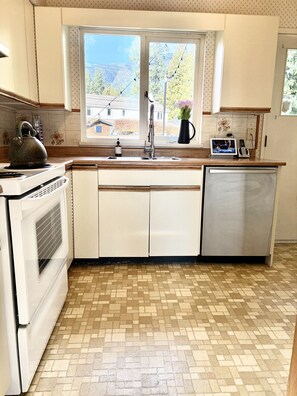 A kitchen with a window, a kettle, a sink, a stove, and a dishwasher. - Mountain Mid-Century Sanctuary: Huge Yard, Views & Adventure at Your Door (North Vancouver)