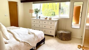 Une chambre à coucher comprenant un lit, une commode, une fenêtre donnant sur des arbres, un miroir et un petit tabouret.