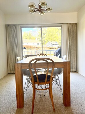 A dining area with a wooden table and chairs, a chandelier, and a view of a residential area through a large window. - Mountain Mid-Century Sanctuary: Huge Yard, Views & Adventure at Your Door (North Vancouver)