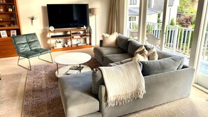Un salon avec un canapé gris, une petite table ronde, un fauteuil en cuir noir et un téléviseur fixé au mur.