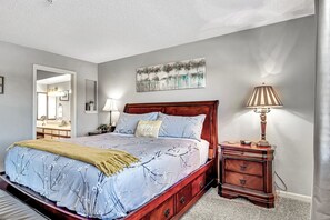 Une chambre à coucher avec un lit en bois, une table de chevet avec une lampe, un tableau accroché au mur, et une salle de bain visible à travers une porte ouverte.