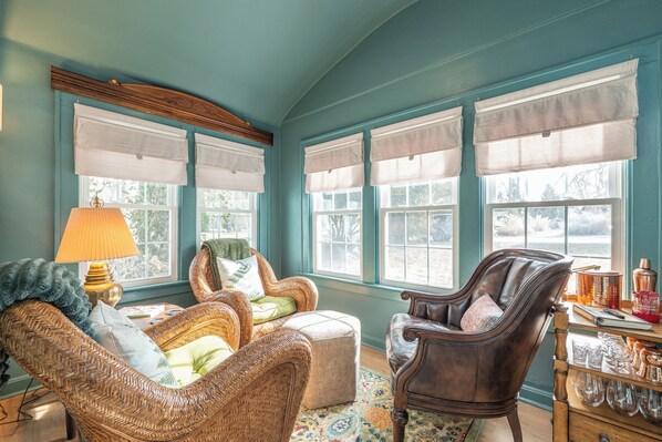 Una habitación acogedora con mobiliario de mimbre, un aparador de madera con cristalería y ventanas grandes con cortinas blancas.