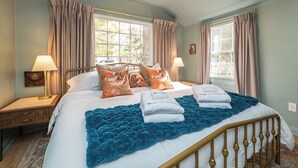 Un dormitorio con una cama, dos lámparas de noche, una ventana con cortinas y una mesita de noche con lámpara.