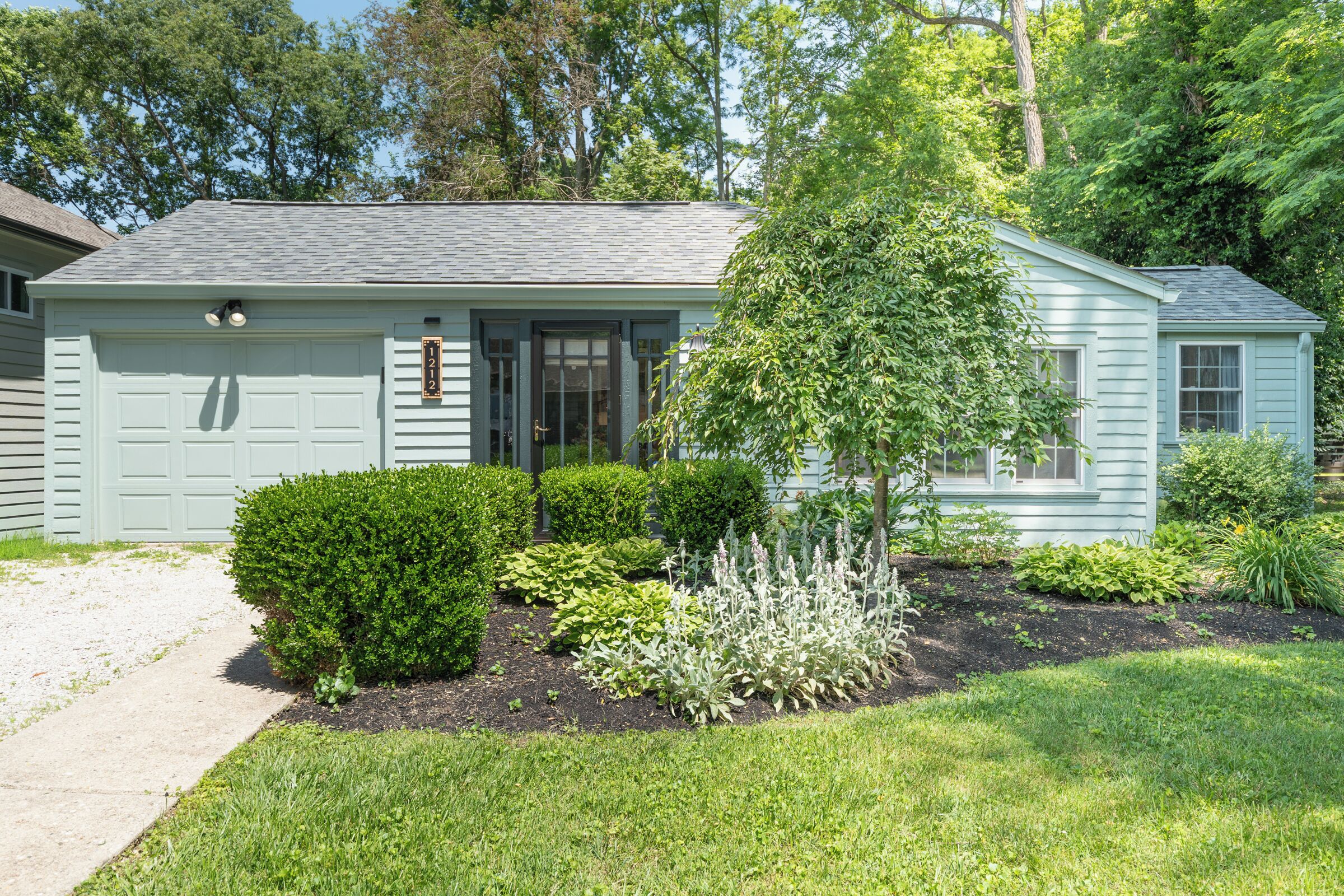A light blue single-story house with a front garage, a small front yard with various shrubs and a tree, and a clear sky.