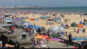 Una playa concurrida, con muchas tiendas de campaña y sombrillas, un muelle al fondo y una turbina eólica.