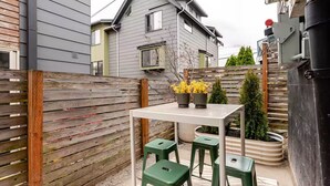 Een klein terras buiten met een tafel en vier groene stoelen, een houten hekwerk en een huis op de achtergrond.