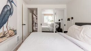 Een slaapkamer met een groot bed, nachtkastje, lamp en een wandplank. Er is een deur naar een andere kamer met een bed en een raam.