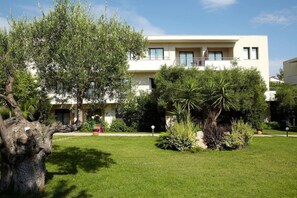 A modern two-story building with a flat roof, a balcony on the upper floor, and a well-maintained garden with various plants and a large tree. - Renaissance Hanioti Resort (Kassandra)