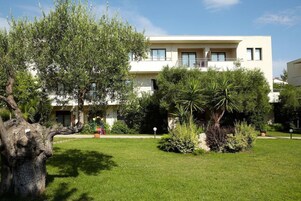 Un edificio moderno de dos pisos con techo plano, un balcón en el nivel superior y un jardín bien cuidado con diversas plantas y un árbol grande.
