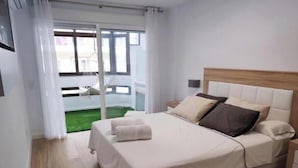Un dormitorio con cama, mesita de noche, ventana con cortinas y vista a un edificio.