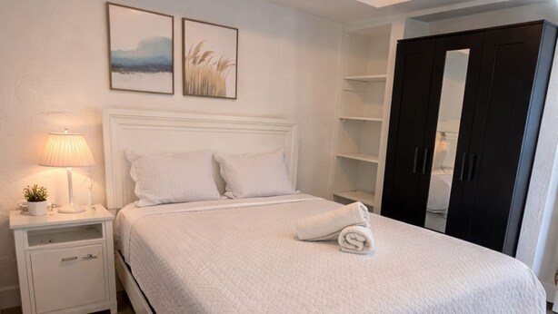 Una cama bien hecha con colcha blanca, una lámpara de noche y una planta pequeña sobre una mesita de noche blanca. Encima de la cama, hay dos cuadros en la pared.