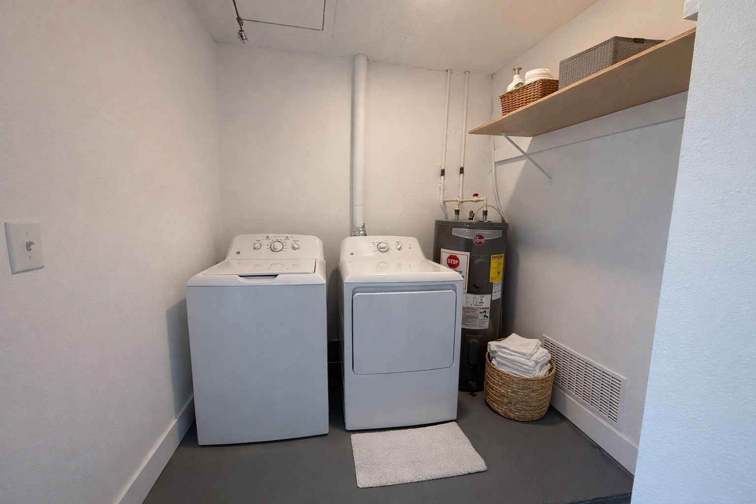 Une buanderie équipée de deux machines à laver à chargement frontal, d’un chauffe-eau et d’un panier en osier contenant des vêtements pliés.