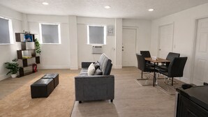 Una sala de estar moderna con un sofá gris, una mesa de comedor con sillas, una estantería y un ventanal grande.