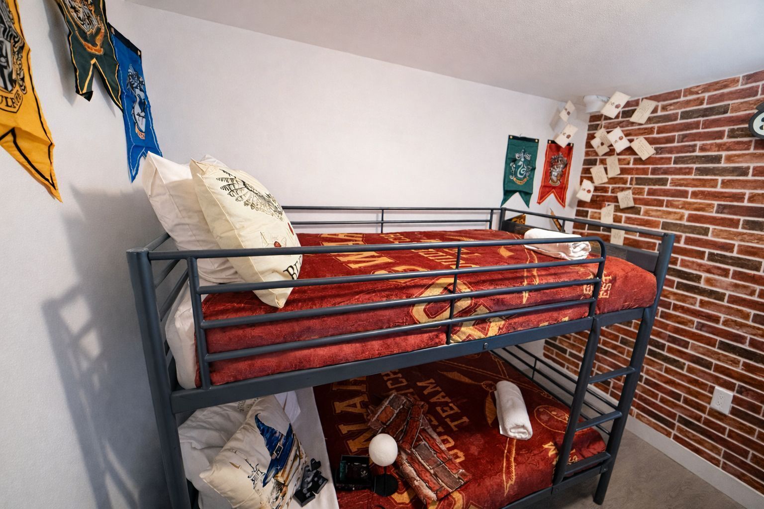 Una cama litera con una colcha estampada roja, almohadas y una manta. Hay banderas en la pared y un adorno de pared de ladrillo.