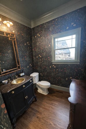 A bathroom with a dark floral wallpaper, a wooden floor, a white toilet, a black vanity with gold handles, and a large mirror. - Upscale 5BR House in Morningside – Perfect for World Cup Groups (Atlanta)