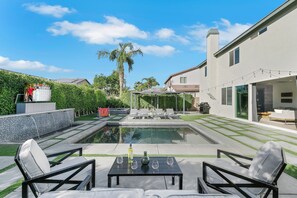 Ein moderner Poolbereich im Freien mit Liegestühlen und einem Tisch, umgeben von einer gepflegten Rasenfläche und einem Haus.