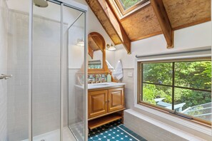 A bathroom with a glass shower enclosure, wooden cabinets, a window with a view of greenery, and a tiled floor.