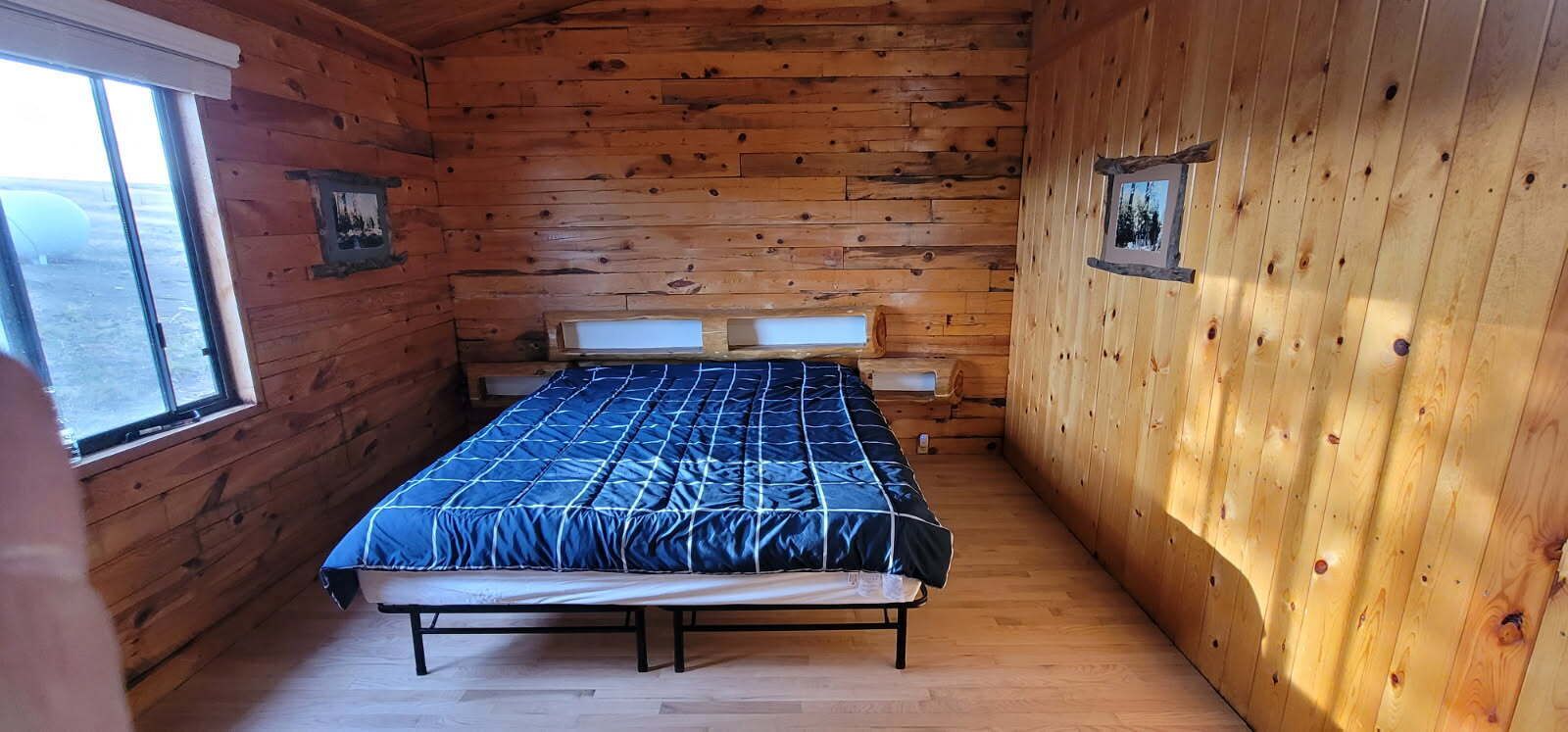 Une petite pièce en bois, aménagée comme une cabane, avec un lit, une fenêtre et deux tableaux encadrés accrochés aux murs.