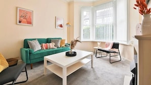 A living room with a green sofa, a white coffee table, and a black chair. There are two windows with white blinds, a framed picture on the wall, and a vase with dried flowers on the table.