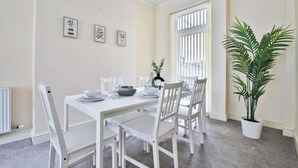 A dining room with a white table set for four, white chairs, a potted plant, and framed artwork on the wall.