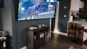 A living room with a large flat-screen TV, a wooden entertainment unit, a kitchen area, and a patterned rug. - Spacious 2-bedroom house in enchanting Kansas City with AC, NSD-STRMAJ-00254  (Kansas City)