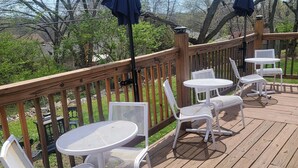A wooden deck with white metal chairs and tables, surrounded by trees and a wooden railing. - Spacious 2-bedroom house in enchanting Kansas City with AC, NSD-STRMAJ-00254  (Kansas City)