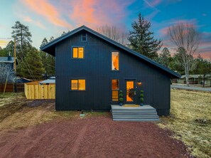Une maison à deux étages, de couleur sombre, avec un toit en pente, un porche à l’avant et une allée menant à l’entrée.