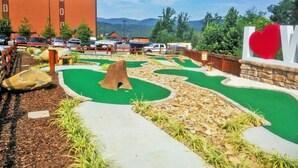 Un campo da mini golf con un manto erboso verde, una base di partenza in legno e un cartello a forma di cuore rosso.