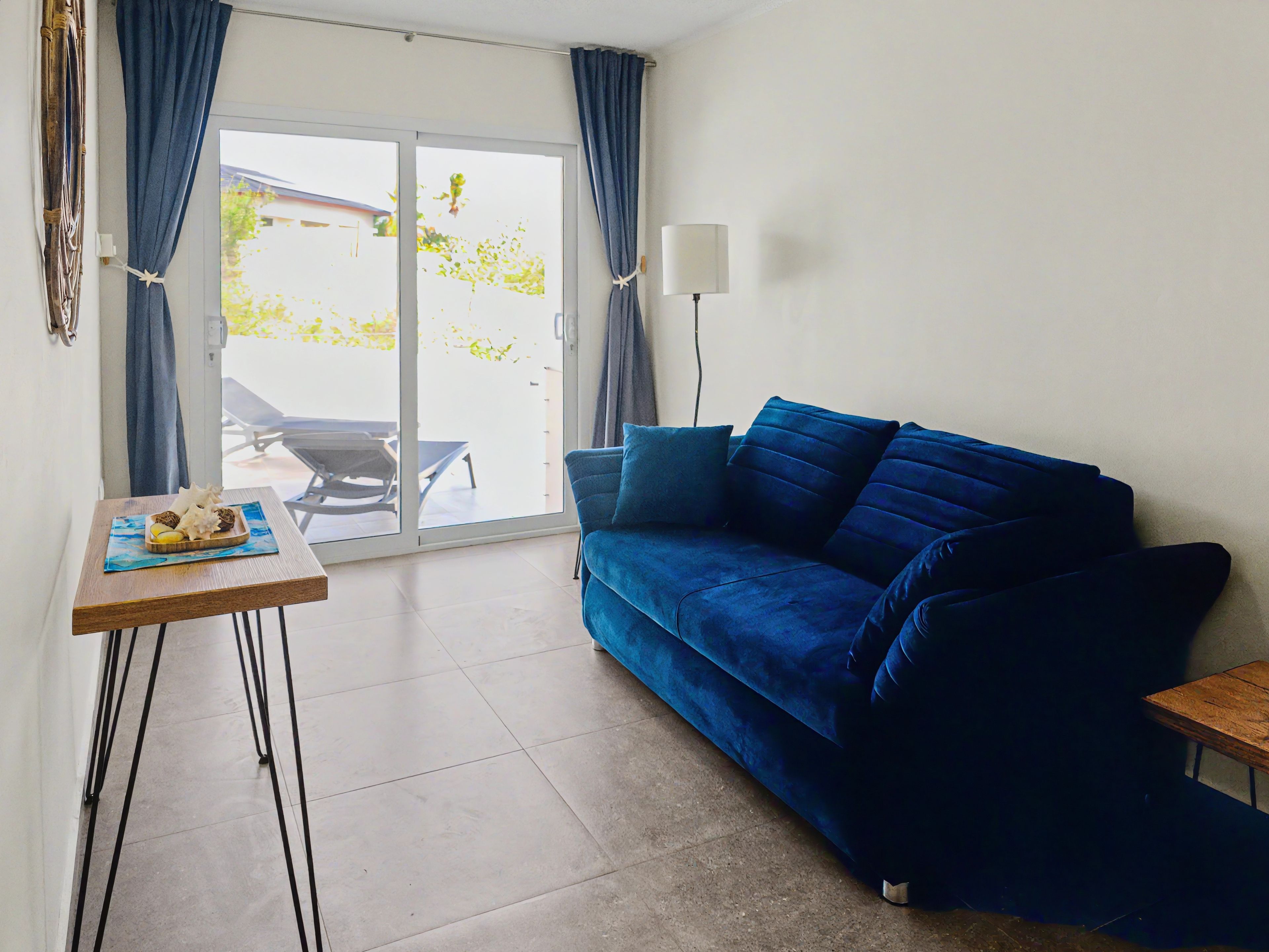 Ein Wohnzimmer mit einem blauen Sofa, einem kleinen Tisch mit einer Dekoration und Blick durch eine Schiebetür.