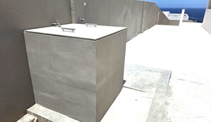 A grey, rectangular outdoor water feature with a faucet and a metal handle, adjacent to a building with a concrete exterior and a view of the sea. - Panoramic 2BR: Ocean, Salt Pans with Flamingos & Infinity Pool (Cas Abao)
