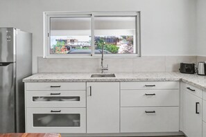 A modern kitchen with a built-in refrigerator, a stainless steel sink, and a window with a view of a garden and buildings. - Panoramic 2BR: Ocean, Salt Pans with Flamingos & Infinity Pool (Cas Abao)