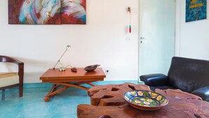 A living room with a wooden coffee table, a black leather sofa, a wooden chair, a table lamp, and abstract paintings on the walls. - Green Oasis Elba- outdoor privacy & walk to the beach- privacy, greenery and the sea (Lacona)