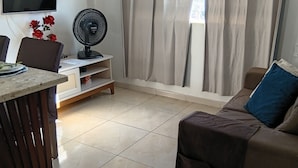 Un salon avec un canapé, un téléviseur fixé au mur, un ventilateur et une fenêtre avec des rideaux.