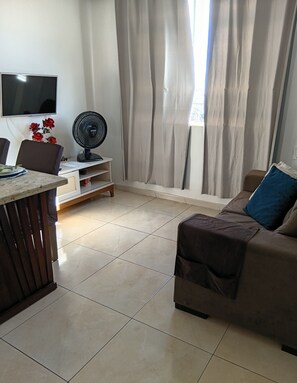 Uma sala de estar com sofá, televisão na parede, ventilador e janela com cortinas.