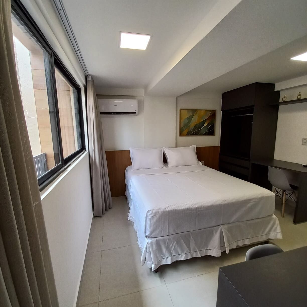 A hotel room with a bed, a desk, a chair, a TV, and an air conditioning unit.