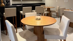 A modern dining area with round wooden tables and upholstered chairs, a kitchen in the background, and a neutral color palette. - Flat Cabo Branco Aurea 50m do mar 403 (João Pessoa)