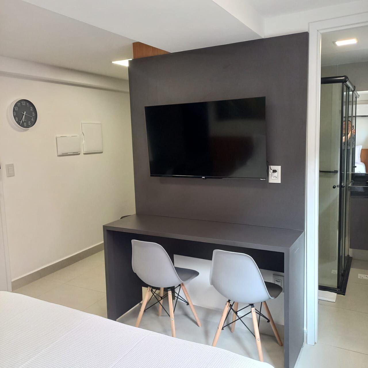 Ein modernes Zimmer mit Flachbildfernseher an einer dunklen Wand, zwei grauen Stühlen, einem kleinen Schreibtisch und einer Wanduhr.