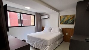 A hotel room with a bed, a desk, a chair, a window with curtains, an air conditioning unit, and a painting on the wall. - Flat Cabo Branco Aurea 50m do mar 403 (João Pessoa)