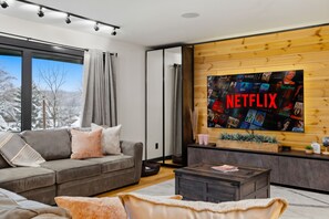 A modern living room with a large flat-screen TV mounted on a wooden wall, a grey sofa with cushions, a coffee table, and a view of a snowy landscape through a large window. - Spa-Style Luxury Lodge w/ Sauna, Hot Tub & Games (Tannersville)