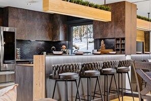 A modern kitchen with dark cabinetry, a stainless steel refrigerator, a wooden bar counter with stools, and a view of a snowy forest through a window. - Spa-Style Luxury Lodge w/ Sauna, Hot Tub & Games (Tannersville)