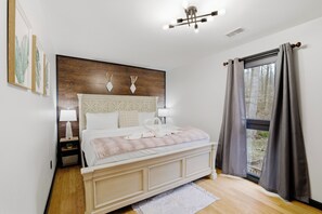 Ein Schlafzimmer mit Bett, Nachttischen, einer Lampe, einem Fenster mit Vorhängen und Blick auf Bäume.