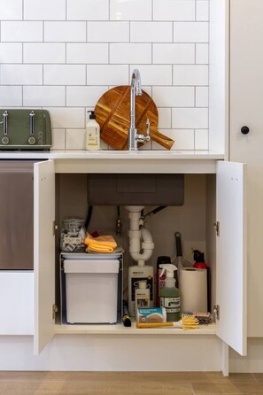 Eine Küche mit weiß gefliestem Spritzschutz, einem Holz Schneidebrett auf der Arbeitsplatte und einem darunterliegenden Schrank mit Reinigungsmitteln und einem Mülleimer.