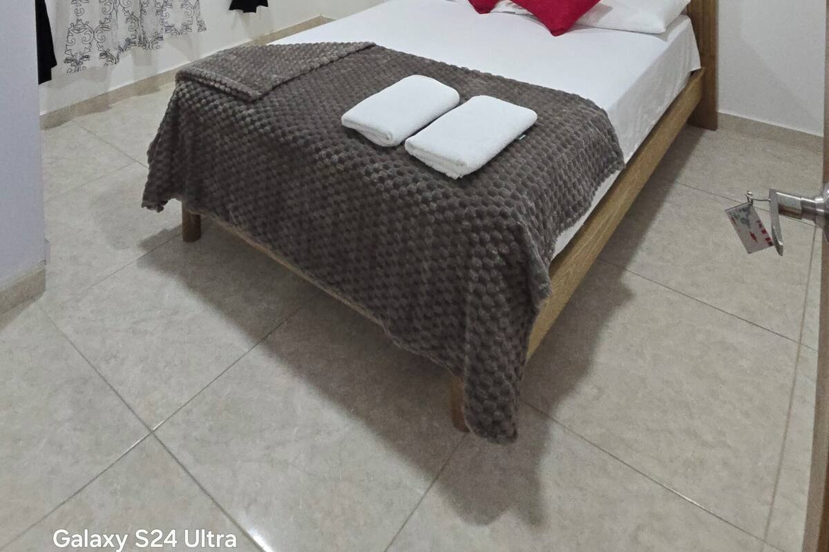 A bedroom with a bed, pillows, a bedside table, a window with curtains, a shelf with decorative items, and a door.